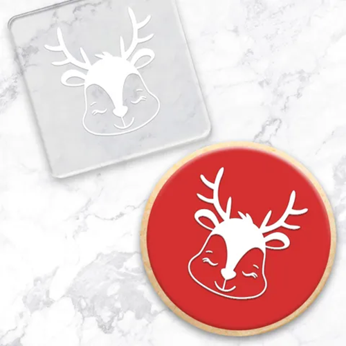 An acrylic debosser with a reindeer head design, shown alongside a sample round cookie cutout with a bright red background and the same reindeer design.