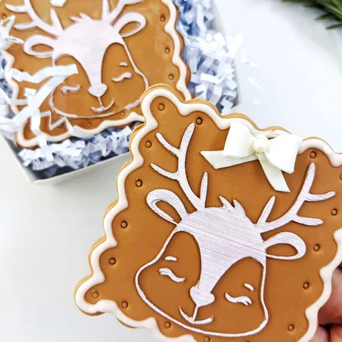 A close-up of a decorated gingerbread cookie shaped like a sleeping reindeer with white icing details, a white bow, and antlers, held in front of another similar cookie in a gift box with white crinkle paper.