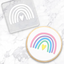 A round white-frosted cookie with a pastel rainbow and small yellow heart design, shown beside a clear square embossing tool  featuring the same rainbow motif on a white marble surface.
