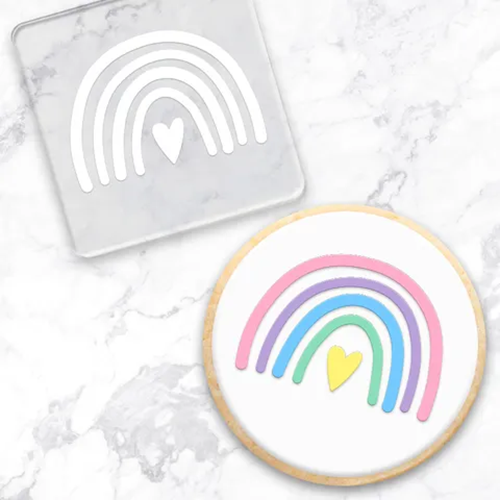 A round white-frosted cookie with a pastel rainbow and small yellow heart design, shown beside a clear square embossing tool  featuring the same rainbow motif on a white marble surface.