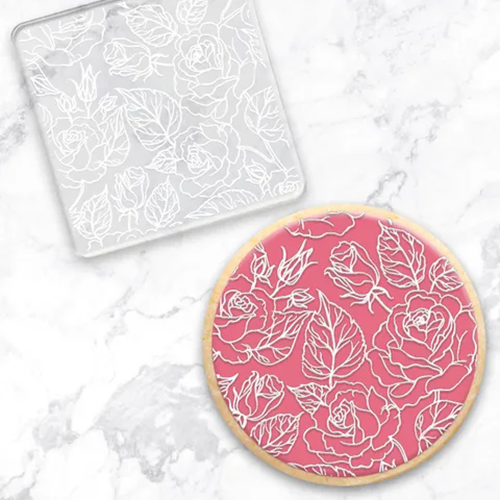 A clear rectangular embossing tool with a white floral rose pattern, shown alongside a round decorated cookie featuring the same white rose design on a pink icing background, set on a white marble surface.