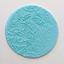 A round turquoise blue fondant icing plaque featuring a raised floral rose pattern, shown on a plain off-white background.