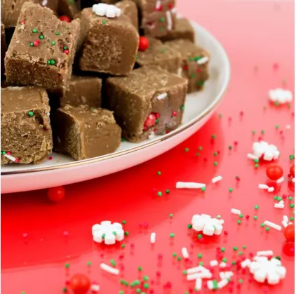 Chocolate fudge cubes decorated with red and green sprinkles, piled on a white plate against a bright red background with scattered festive sprinkles.