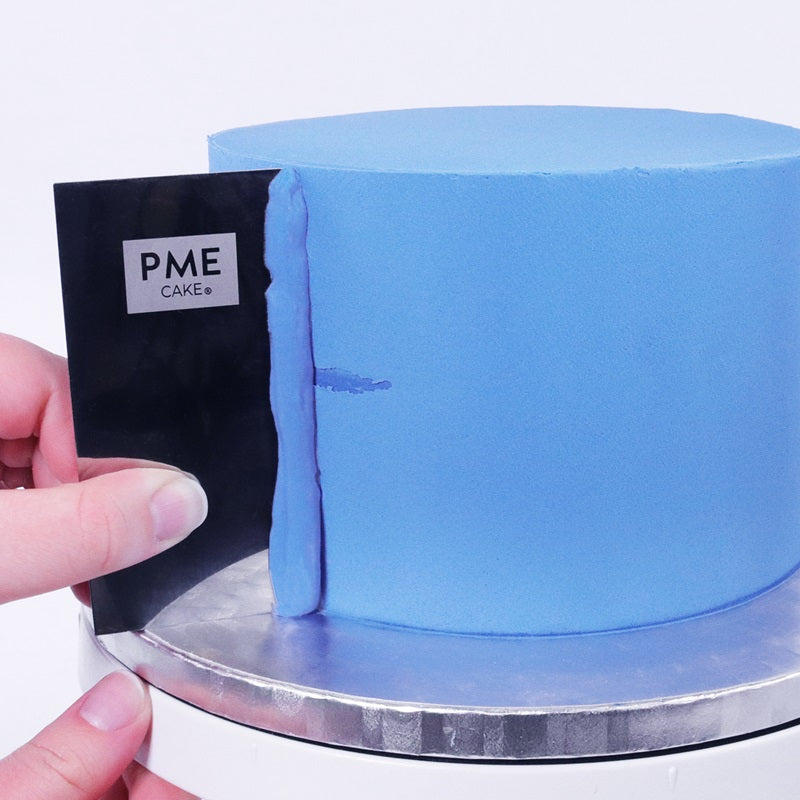 A blue iced cake with a PME metal scraper, that is scraping the side