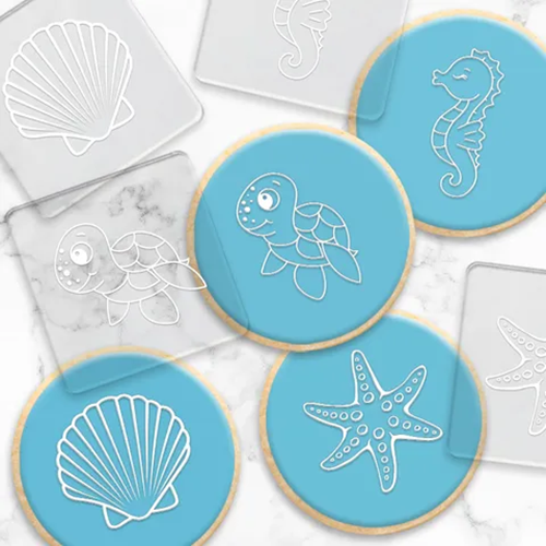 Round sugar cookies decorated with light blue icing and white sea-themed line drawings, including a turtle, seahorse, starfish, and shell, arranged on a white marble surface with clear acrylic embossing tools visible around them.
