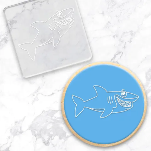 A round blue cookie with a white shark outline, shown beside a clear square acrylic embossing tool featuring the same shark design, on a light marble surface.