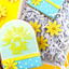 Decorated sugar cookies in a gift box with shredded paper. The front cookie is shaped like an arch with a yellow sun design and the words 'shine bright' in yellow icing.