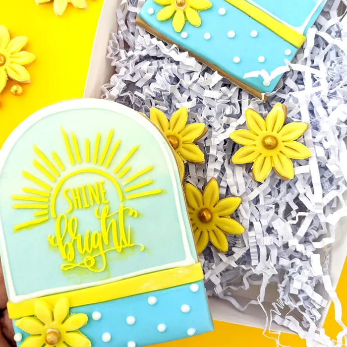 Decorated sugar cookies in a gift box with shredded paper. The front cookie is shaped like an arch with a yellow sun design and the words 'shine bright' in yellow icing.