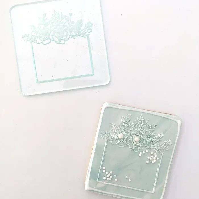 A clear debosser with a floral square frame design placed on a whitesurface, with an example imprint on a sage cookie below it.