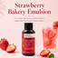 A bottle of LorAnn Strawberry Bakery Emulsion centered on a soft pink background, with large red headline text above. Fresh strawberries and a pink strawberry drink are arranged around the bottle as styled props.