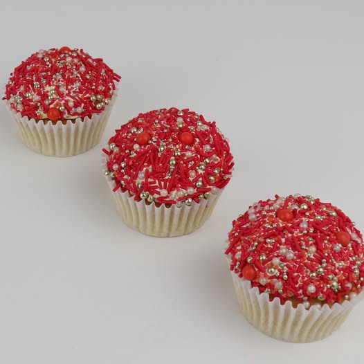 Three cupcakes in white paper liners topped with red frosting and sprinkles, arranged diagonally on a light gray background.