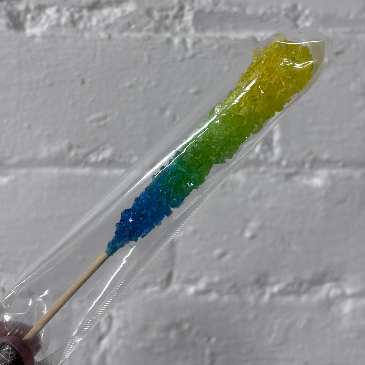 A rainbow-colored rock candy stick in a clear plastic sleeve, resting against a white brick wall background.