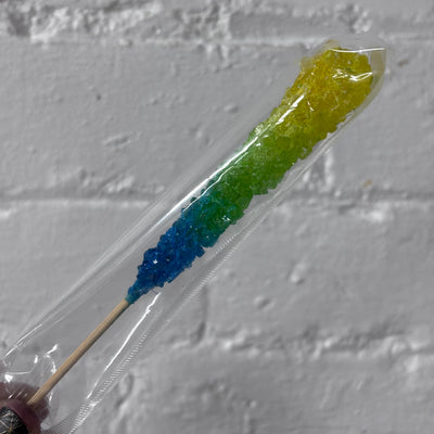 A rainbow-colored rock candy stick in a clear plastic sleeve, resting against a white brick wall background.