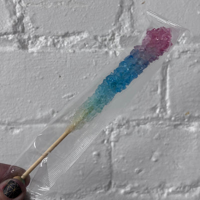 A hand holding a packaged rock candy stick with a pink-to-blue gradient, wrapped in clear plastic against a white brick wall background.