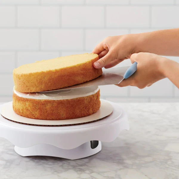 Hands holding a Wilton cake lifter. Lowering a layer of cake down on top of a second layer, sitting on a cake turntable