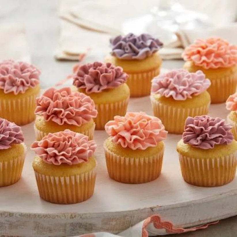 A group of yellow cupcakes in paper liners, decorated with ruffled piped frosting in pink, peach, and lavender, arranged on a rustic white wooden board.