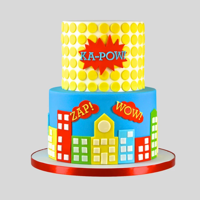 A two-tier superhero-themed birthday cake with yellow polka dots on the top tier, a red comic-style burst with 'KA-POW!' in blue, and colorful fondant buildings on the bottom tier with 'ZAP!', 'WOW!', and 'YUP!' text.