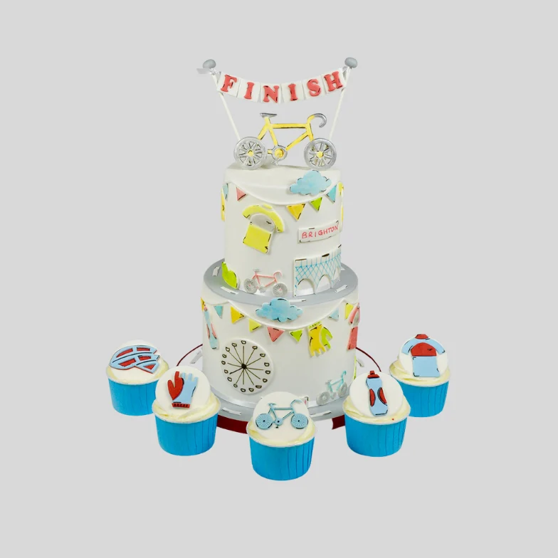 A 3-tier white cake decorated with colorful bicycle-themed illustrations, a bicycle topper, and a 'FINISH' banner, surrounded by six blue cupcake toppers on a light gray background.