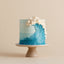 A cylindrical cake decorated with a blue wave design and white piped frosting curls, displayed on a beige ceramic pedestal stand against a solid beige background.