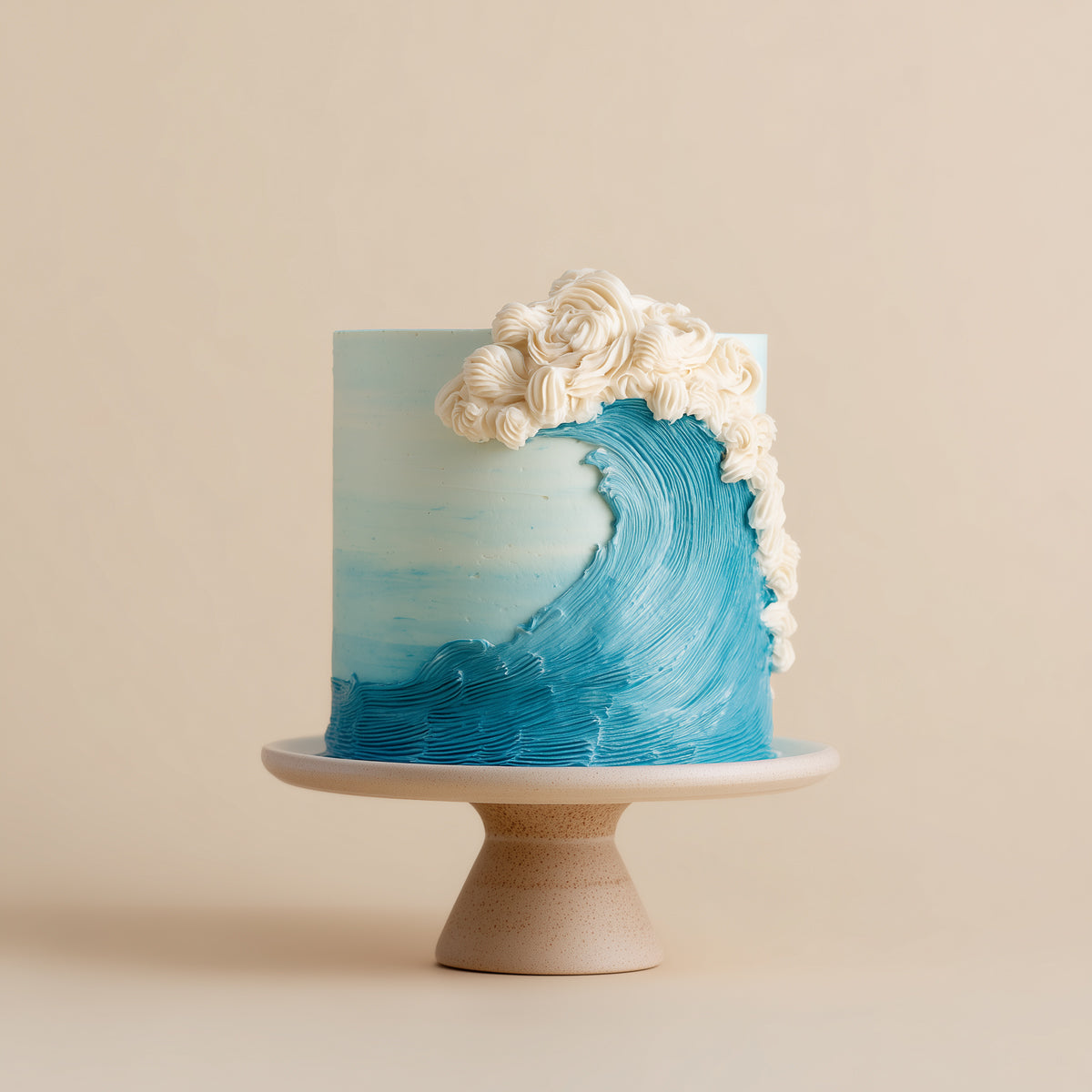 A cylindrical cake decorated with a blue wave design and white piped frosting curls, displayed on a beige ceramic pedestal stand against a solid beige background.