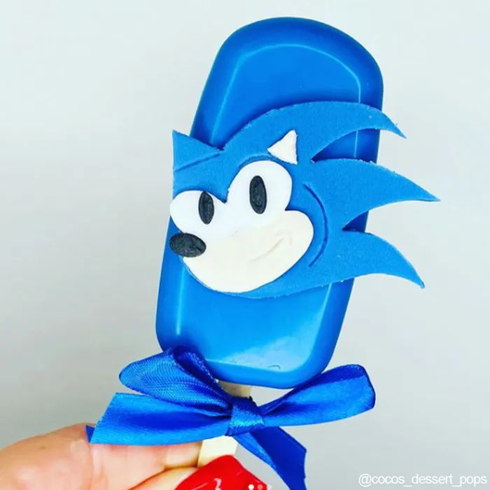 Blue lollipop shaped like a character with a white face and black eyes, held by a hand against a light gray background.