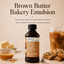 A bottle of LorAnn Brown Butter Bakery Emulsion centered on a warm beige background, with large headline text above. A small bowl of emulsion, butter cubes, a glass of iced coffee with cream swirl, and chocolate chip cookies are arranged around the bottle as styled props.