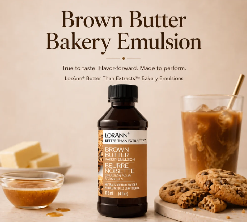 A bottle of LorAnn Brown Butter Bakery Emulsion centered on a warm beige background, with large headline text above. A small bowl of emulsion, butter cubes, a glass of iced coffee with cream swirl, and chocolate chip cookies are arranged around the bottle as styled props.