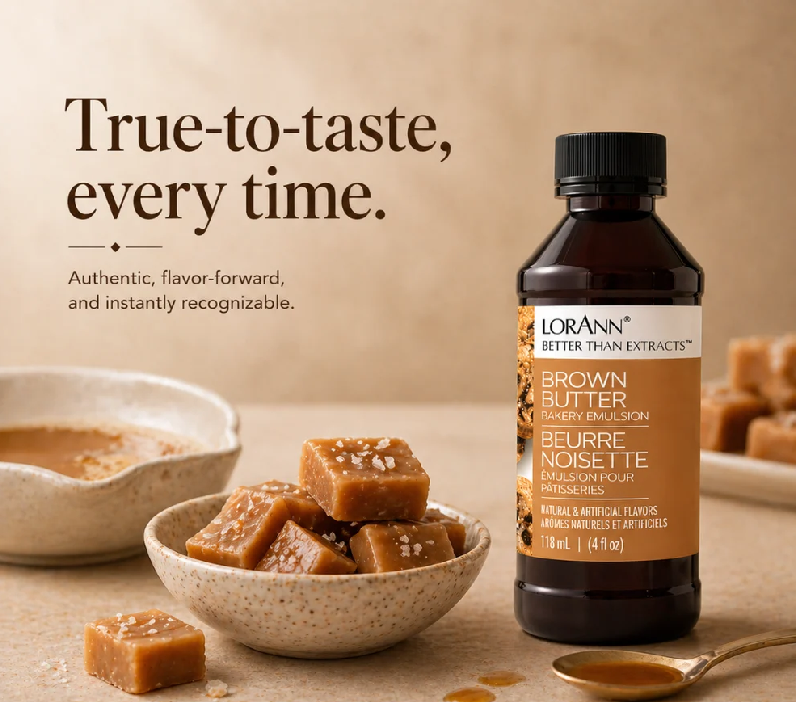 A bottle of Lorann Better Than Extracts Brown Butter flavoring stands on a beige surface beside bowls of caramel-colored candy or sauce, with promotional text reading 'True-to-taste, every time.'.