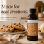 A bottle of LorAnn Better Than Extracts Brown Butter flavor food flavouring emulsion stands on a countertop beside chocolate chip cookies, a small bowl of caramel sauce, and pastries in a warm kitchen setting. Large text on the left reads, 'Made for real creations.