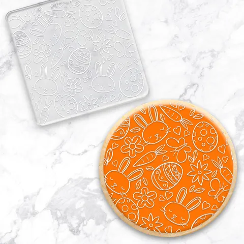 Debosser with Easter design along with a finished cookie on an orange round fondant circle on a white marble surface