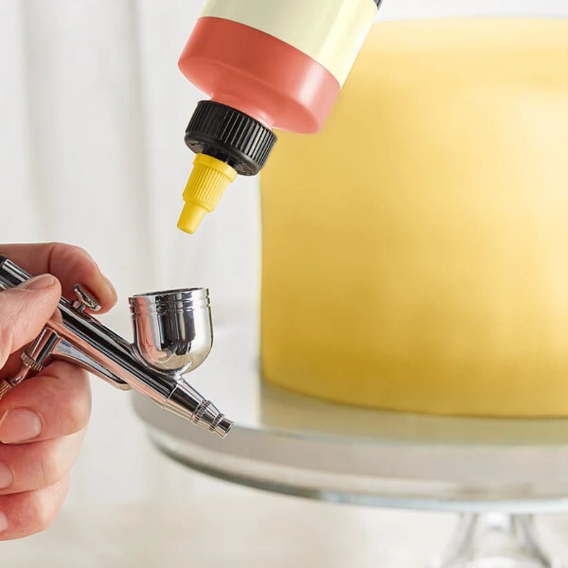 A close-up of an airbrush applicator being used to apply a peach-colored icing or paint onto a yellow frosted cake on a glass cake stand.