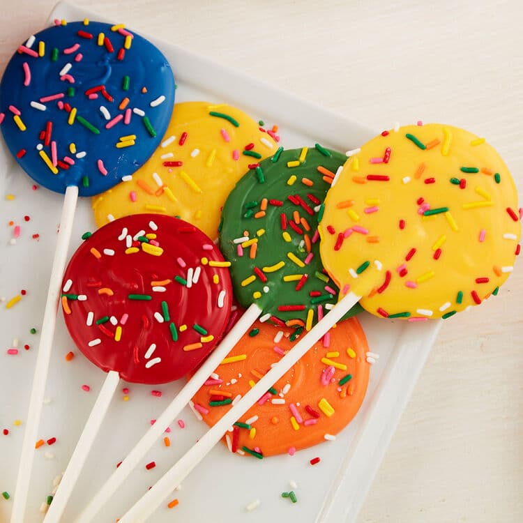 Six round lollipop-style treats coated in bright colors—blue, yellow, green, red, orange, and yellow—each topped with multicolored sprinkles, arranged on a white tray.