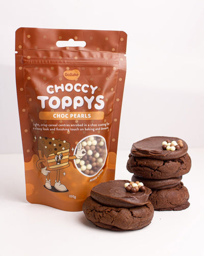 A stand-up pouch of Go bake Choccy Toppys (choc pearls) next to three chocolate chip cookies topped with the white and beige chocolate pearls, on a clean white background.
