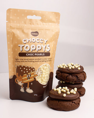 A 100g bag of GoBake Choccy Toppys (choc pearls) stands next to three chocolate cookies topped with white chocolate pearls, set against a clean white background.