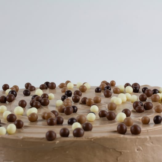 A close-up of a brown frosted cake topped with scattered chocolate and white chocolate candies against a light gray background.
