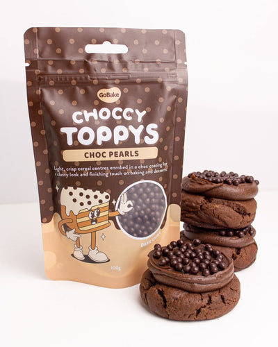 A 100g stand-up pouch of GoBake Choccy Toppys Choc Pearsls next to three stacks of chocolate chip cookies on a white background.