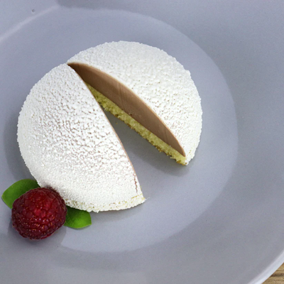 Layered dessert sprayed with white velvet cocoa butter on a gray plate, garnished with a raspberry and mint leaves.
