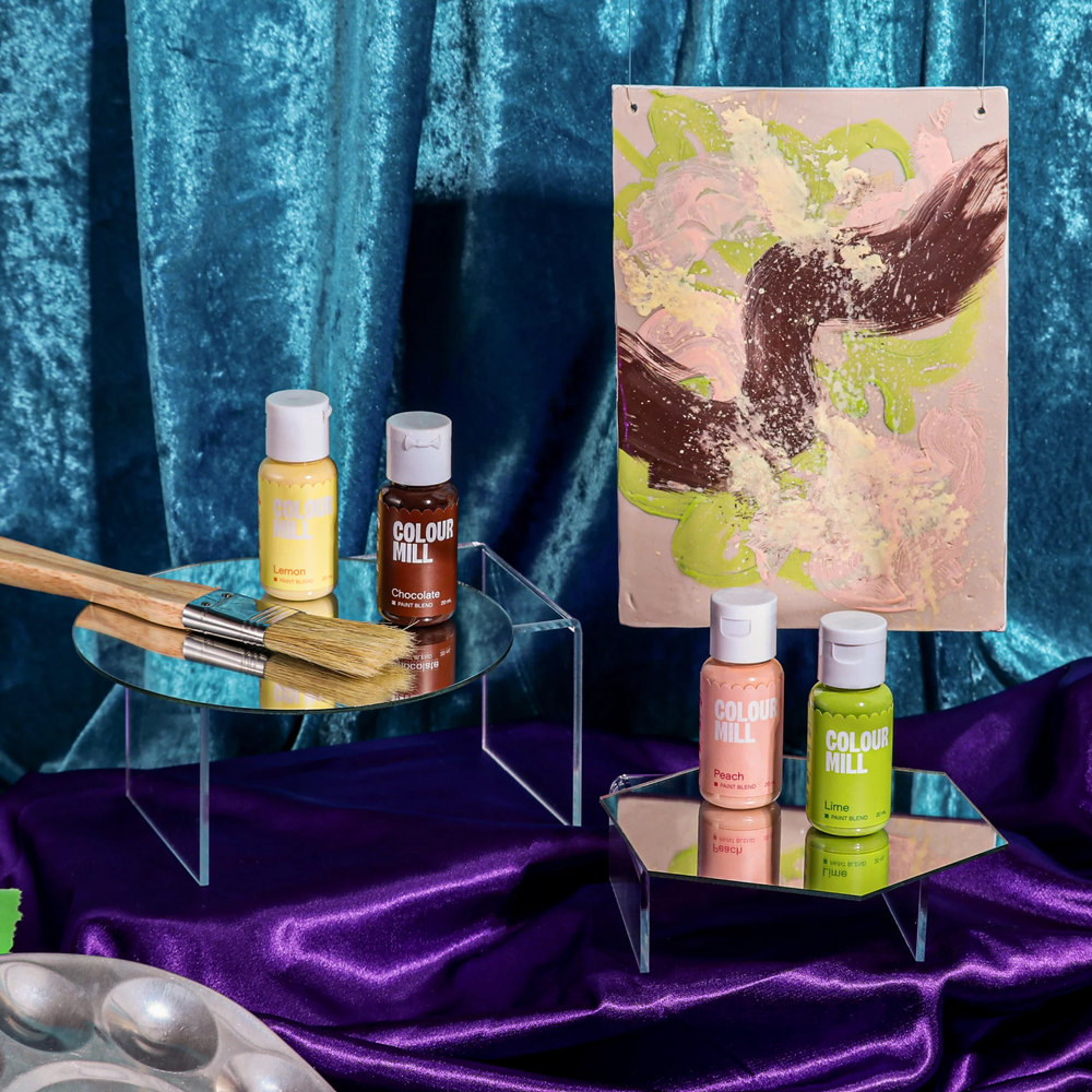 Colorful Colour Mill paint blend bottles displayed on shelves with a blue and purple draped background.