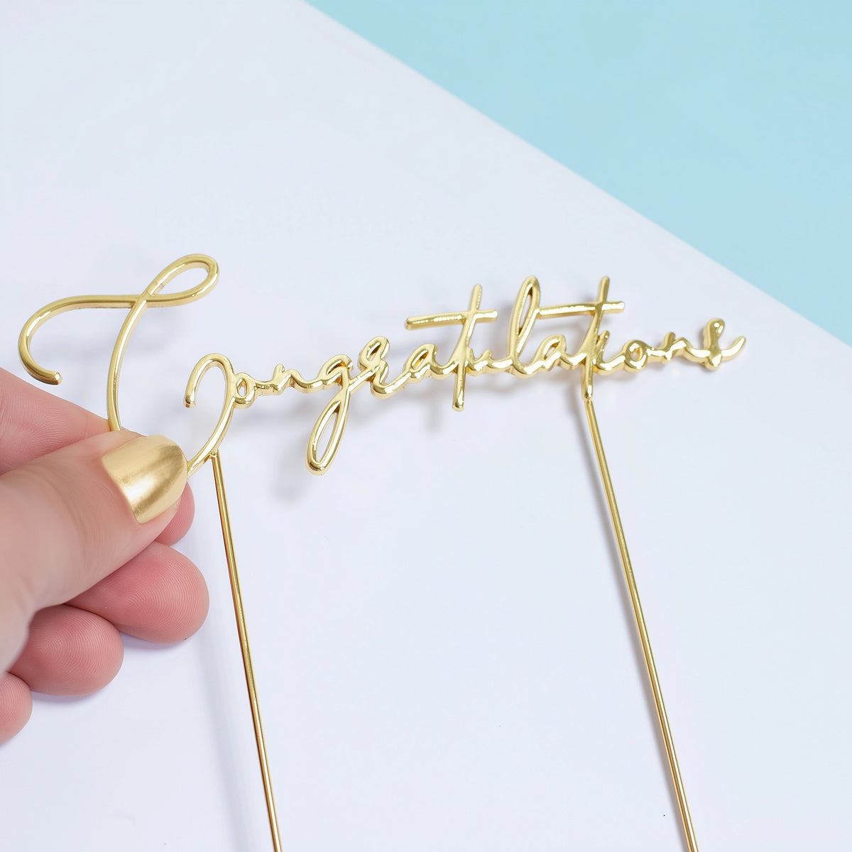 Gold 'Congratulations' cake topper held by a hand on a light blue and white background
