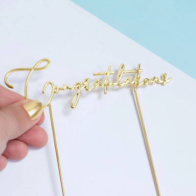Gold 'Congratulations' cake topper held by a hand on a light blue and white background