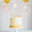 White cake with gold accents and 'Congratulations' topper in front of white balloons and string lights.