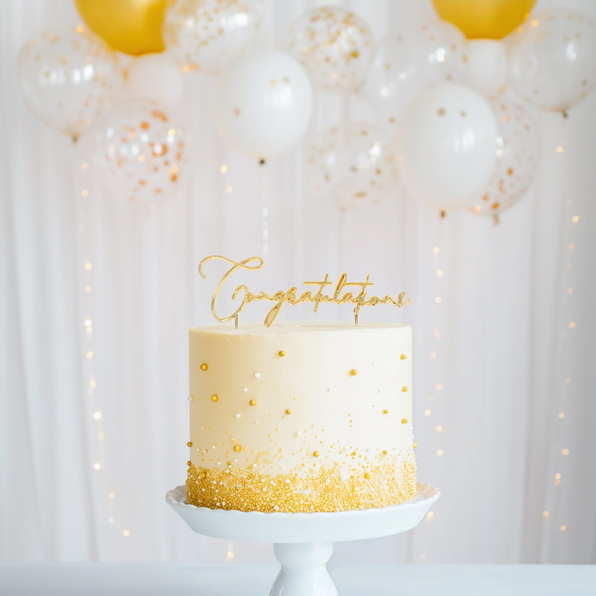 White cake with gold accents and 'Congratulations' topper in front of white balloons and string lights.