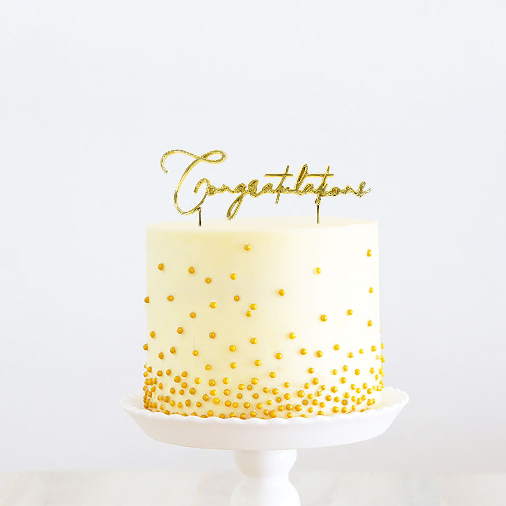 White cake with gold polka dots and 'Congratulations' topper on a white background