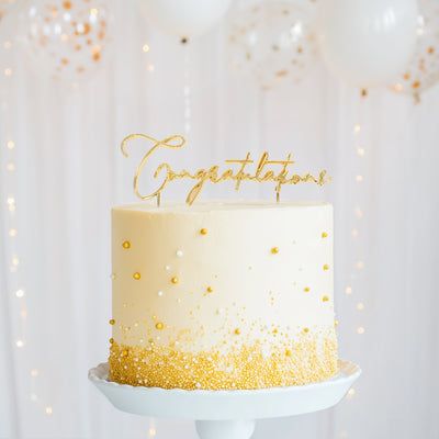 White cake with gold accents and 'Congratulations' topper against a light background