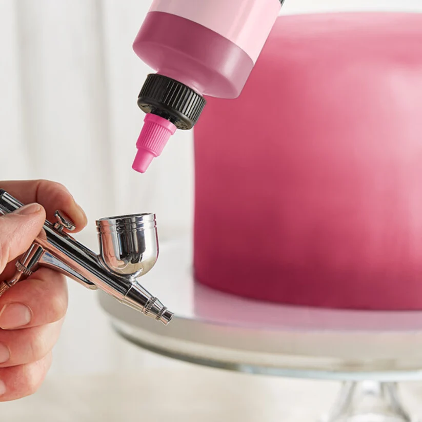 A close-up of a hand holding an airbrush tool while a pink bottle with a nozzle applies pink paint to a round cake on a stand.