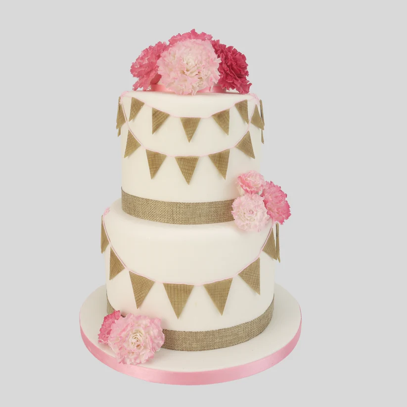 A two-tier white cake decorated with pink and red peony flowers, gold bunting, and burlap-style bands, set against a solid light gray background.