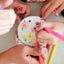 Several hands holding colorful markers around a round white cake decorated with painted flowers, photographed from above on a light tabletop.