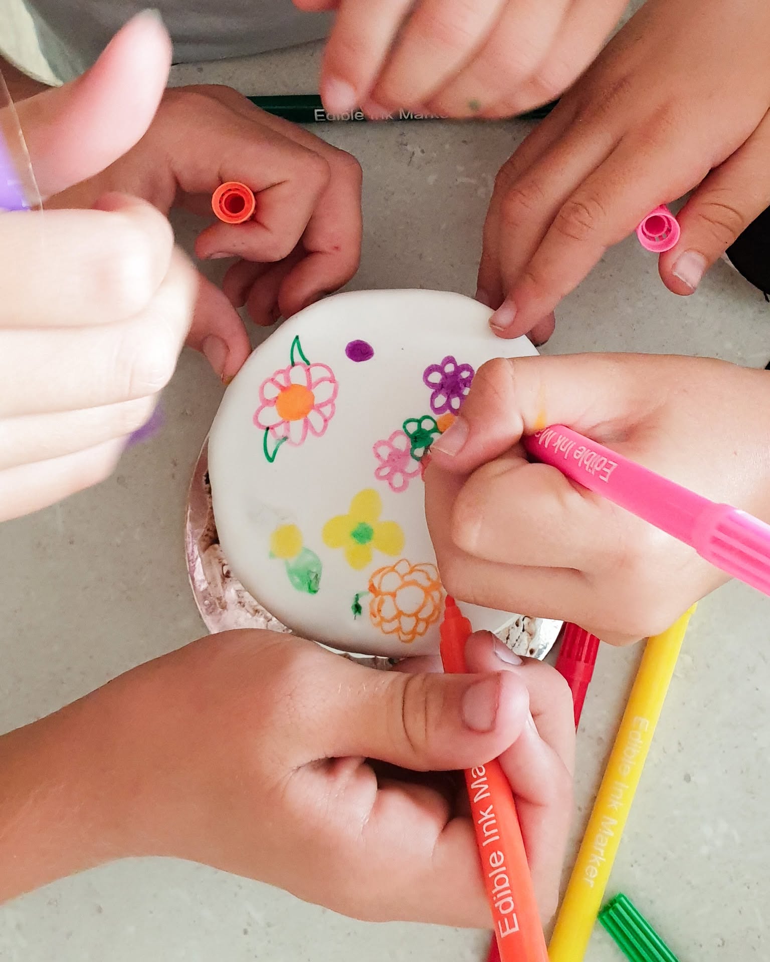 Several hands holding colorful markers around a round white cake decorated with painted flowers, photographed from above on a light tabletop.
