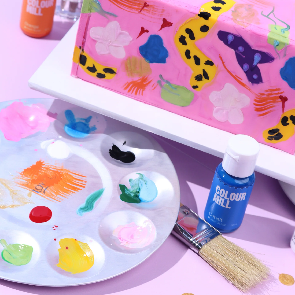 Colorful cake art supplies including a paint palette, brush, and Colour Mill product on a pink background