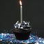 A black frosted cupcake with a single lit silver candle, topped with white, blue, and purple sprinkles, set against a dark background with sprinkles scattered around it.